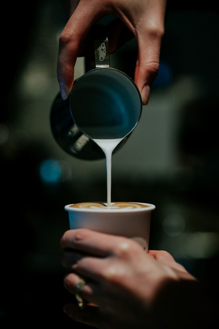 A barista pours steamed milk into a small cup of espresso on a wooden counter with natural light.
