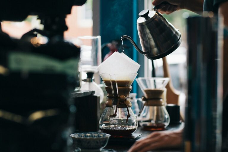 A person measures ground coffee and hot water beside a clean brewer on a kitchen counter.