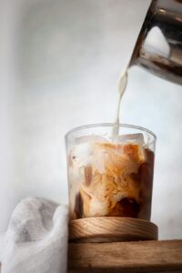 A person pouring coffee concentrate over ice in a clear glass on a kitchen counter.